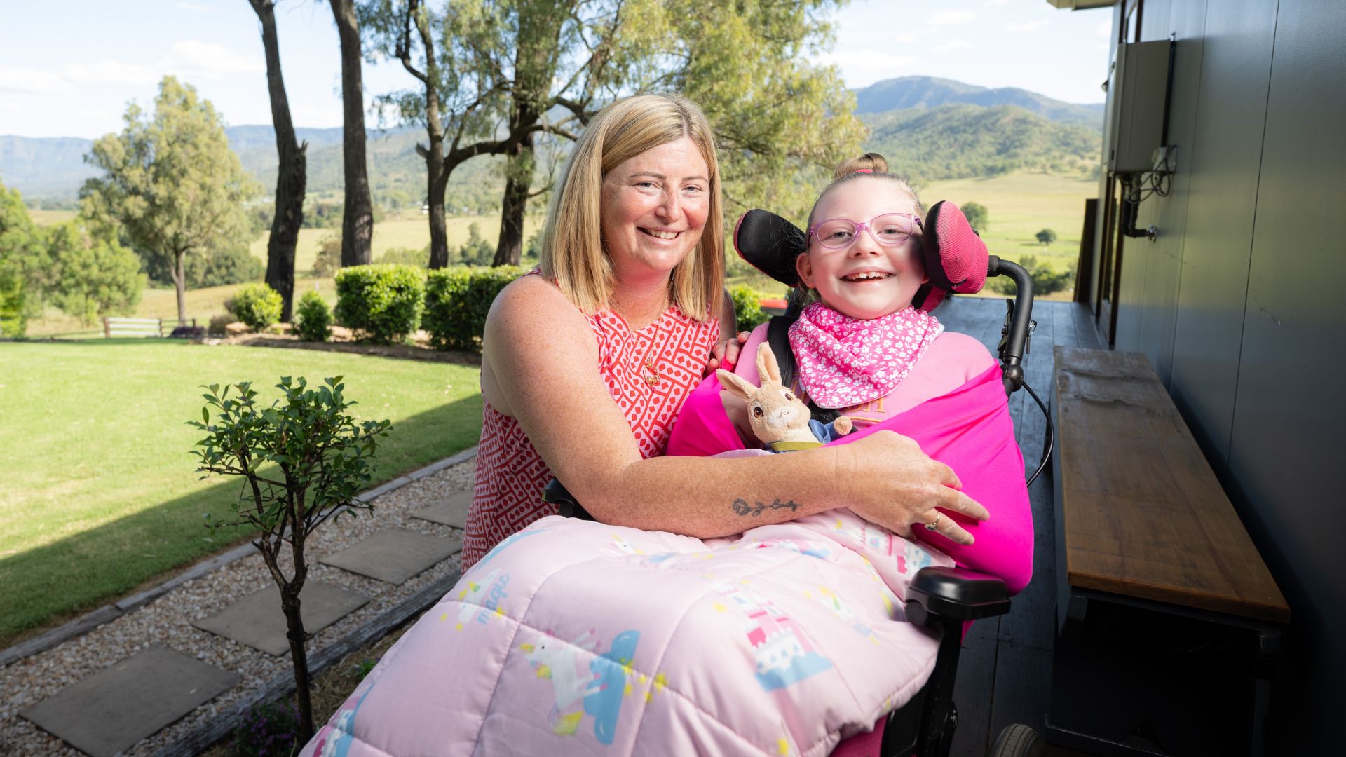 Jessica gently holds Emily, who is wrapped in a pink blanket and seated in a wheelchair with pink head supports. A soft toy rabbit rests on Emily’s lap. The scene is set outdoors with lush green lawns and mountains in the distance, highlighting love and care in disability support.