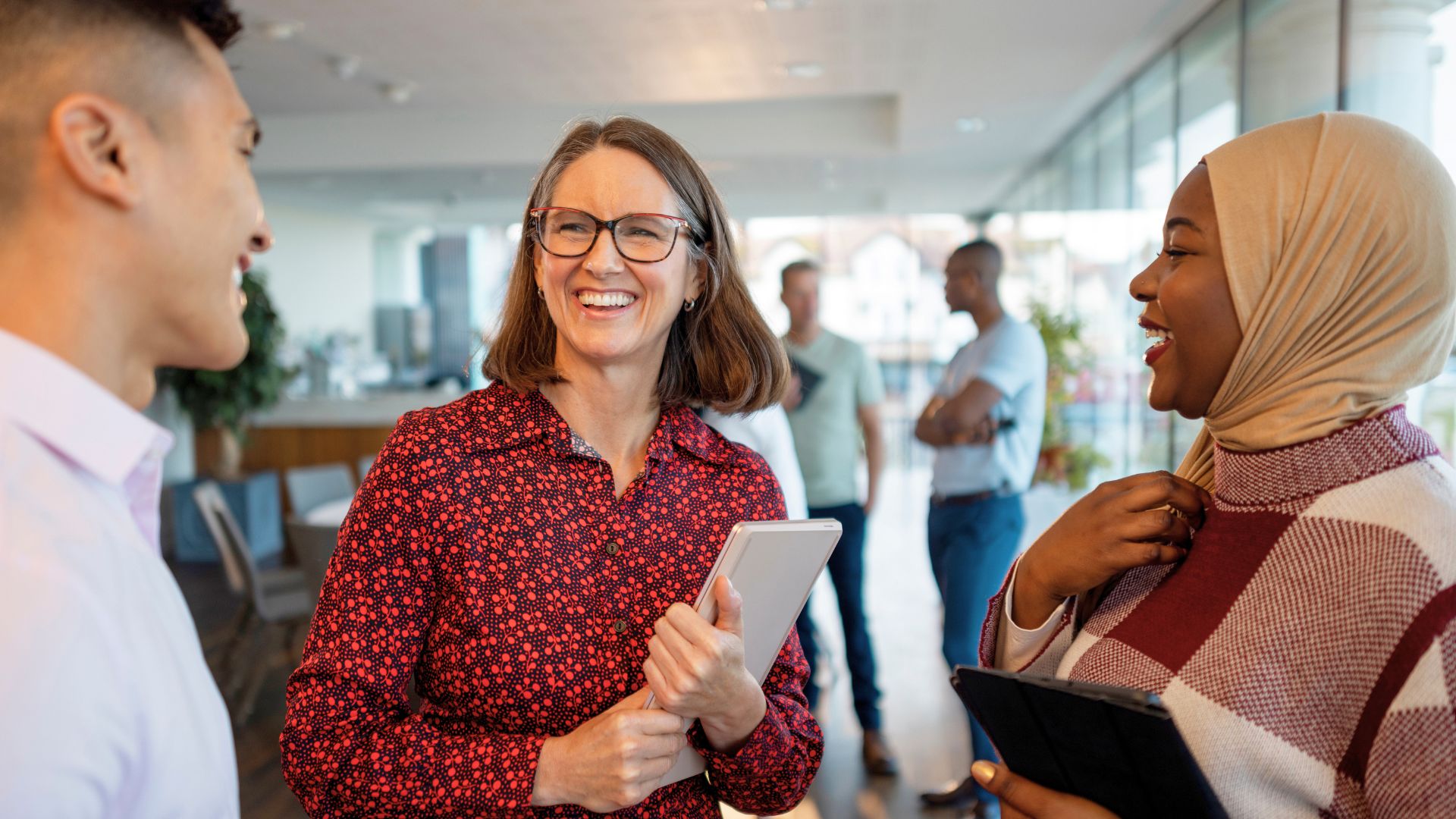 Three colleagues smiling and talking together in a modern office, holding tablets and notepads.