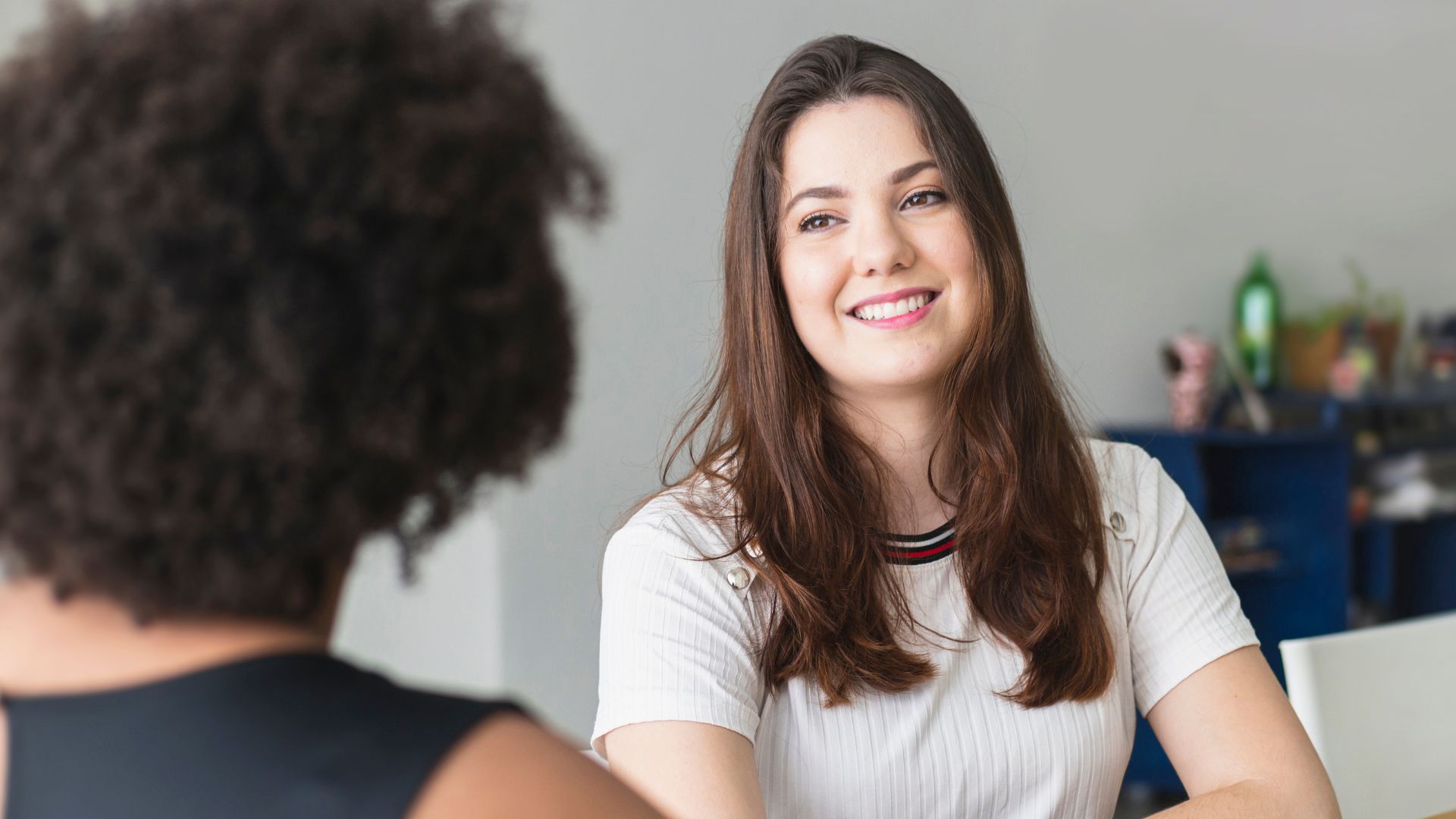 A young woman in a striped shirt sitting in a library or study area, smiling as she talks to another person across the table.