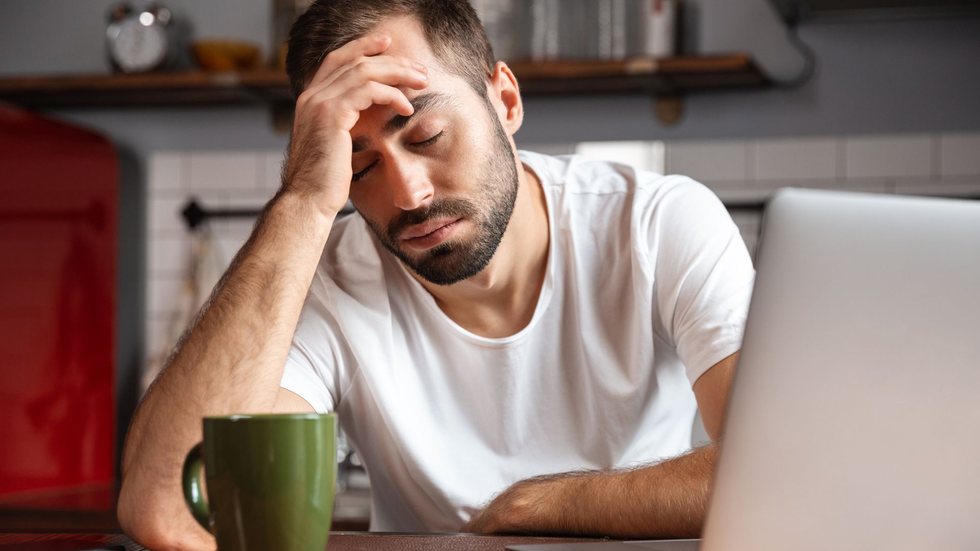 Person leaning on hand while sitting at a table with a laptop and coffee mug, showing fatigue and low energy as common signs of immunodeficiency.