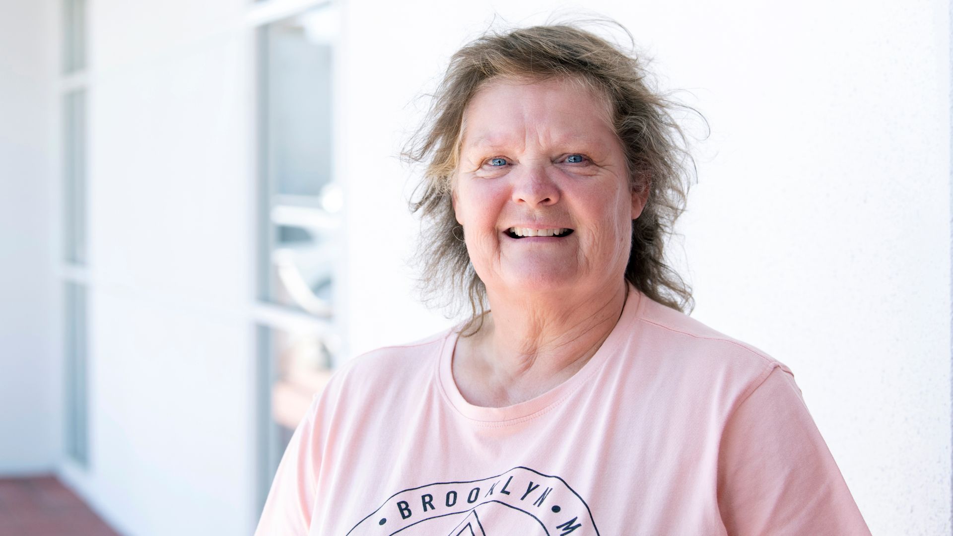 Smiling woman standing outdoors near a building, looking at the camera in a bright setting.