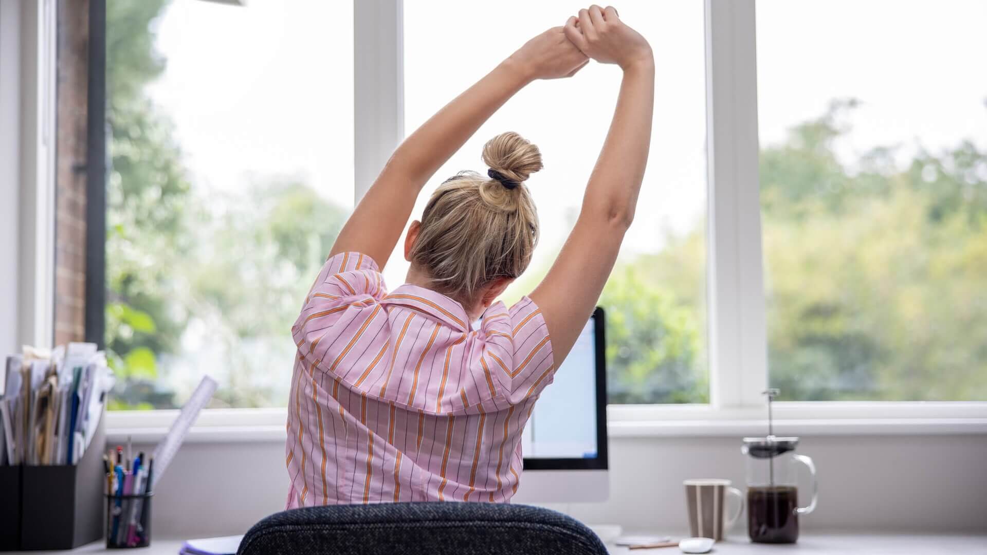 A woman sitting at a desk with arms raised and stretching her back