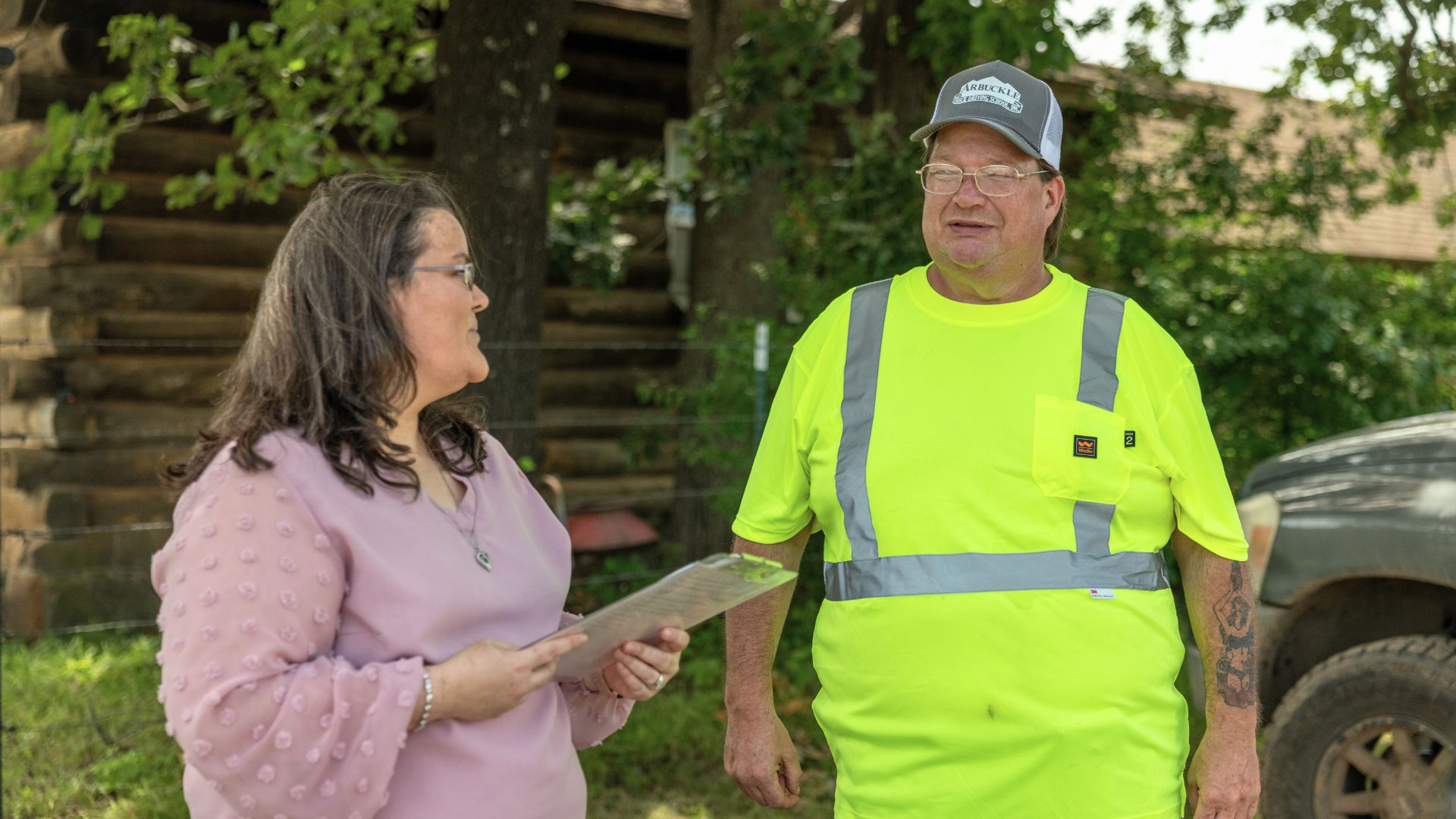Candice wearing a pink blouse and holding papers, and Ocie in a high-visibility yellow vest with a "Caterpillar" cap, engaging in conversation outdoors near a wooden structure and a vehicle.