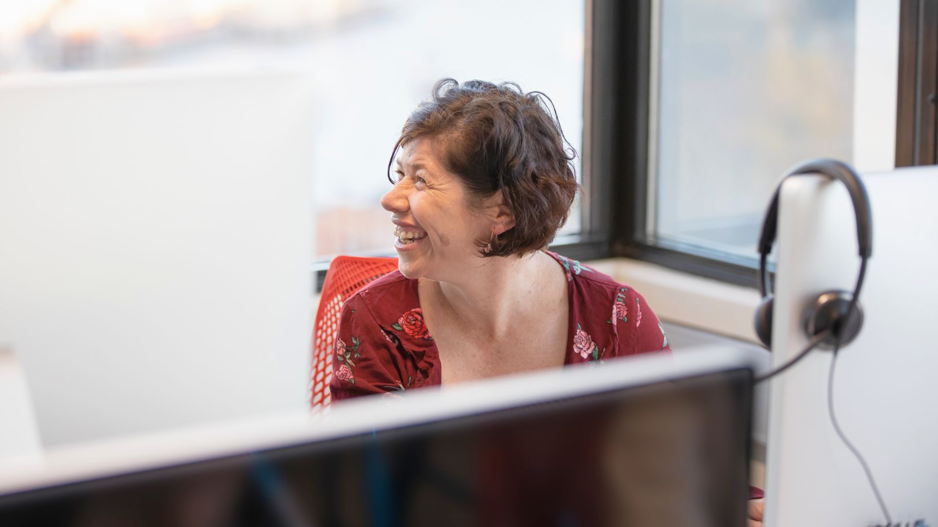 Smiling woman working at a computer in a bright office, looking engaged and supported in a workplace environment.