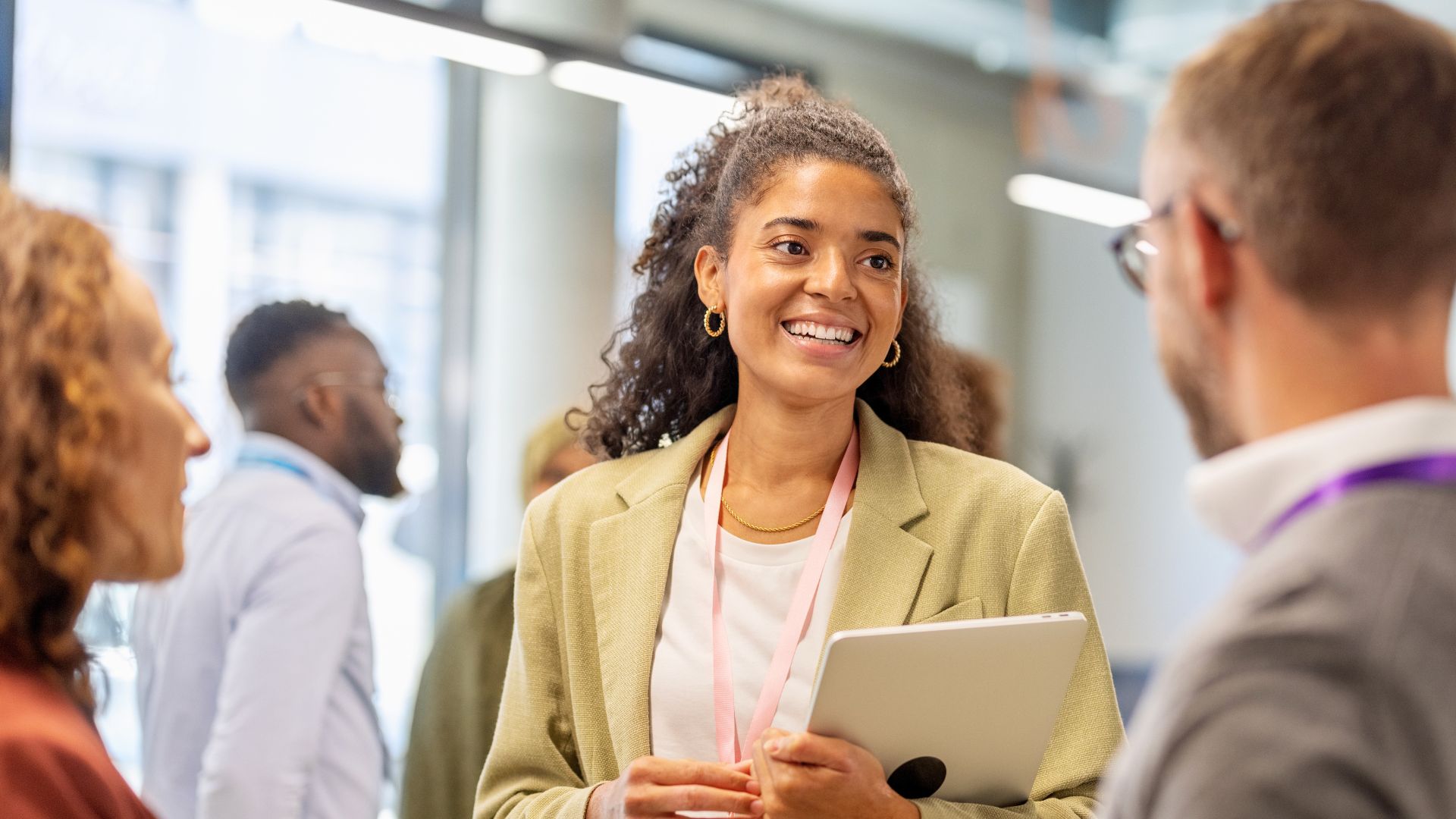 Three professionals chatting in a modern office, with a smiling woman holding a tablet while speaking with colleagues.