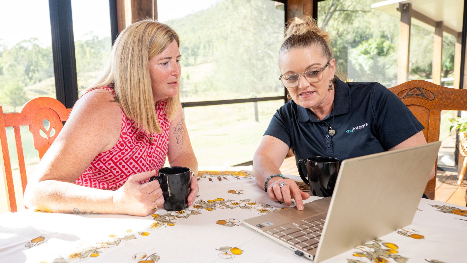 Jessica and a MyIntegra support coordinator sit at a wooden table with coffee mugs, reviewing information on a laptop. The setting is a bright, open space with scenic views outside, representing collaboration in managing complex NDIS plans.