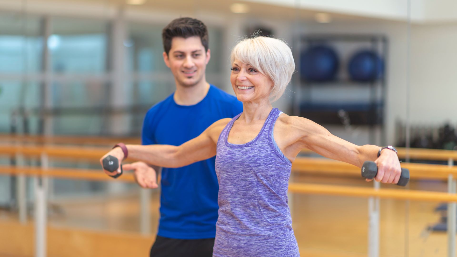 An older woman lifts weights and smiles next to a personal trainer