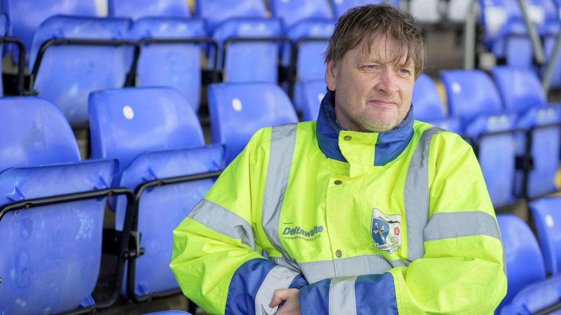 Mark, a steward supported by Ingeus, seated among blue stadium seats at Barrow AFC wearing a high-visibility jacket, representing new employment opportunities through the Pioneer programme.
