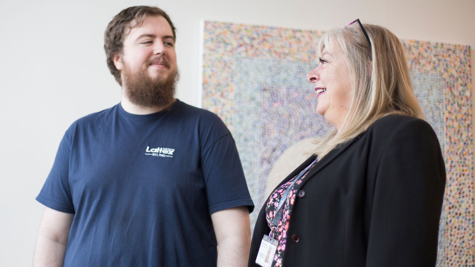 Fraser standing indoors with another person near a colourful wall display, symbolizing his successful transition into employment with Ingeus UK.