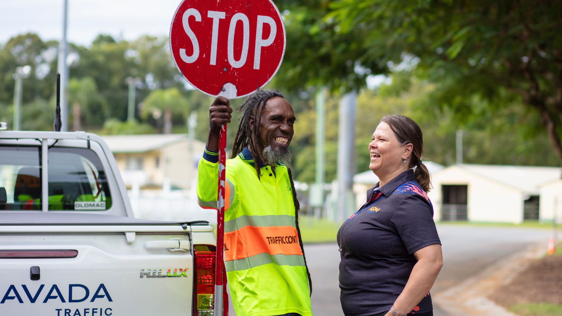 Workforce Australia participant Adam holds a stop sign while standing next to his APM employment consultant Janeah in a First Nations artwork top, both smiling outdoors near a traffic control vehicle.