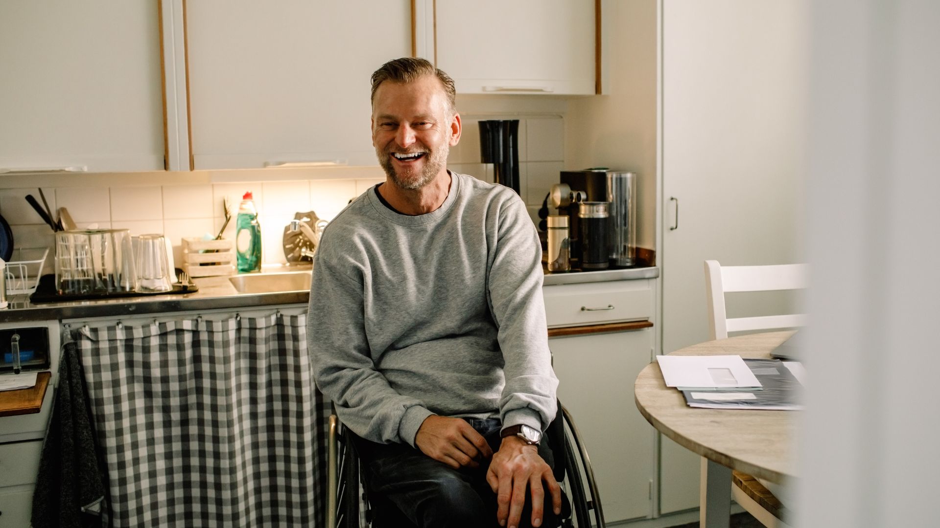 A man using a wheelchair sits in a kitchen, smiling warmly, with household items and a dining table visible around him.