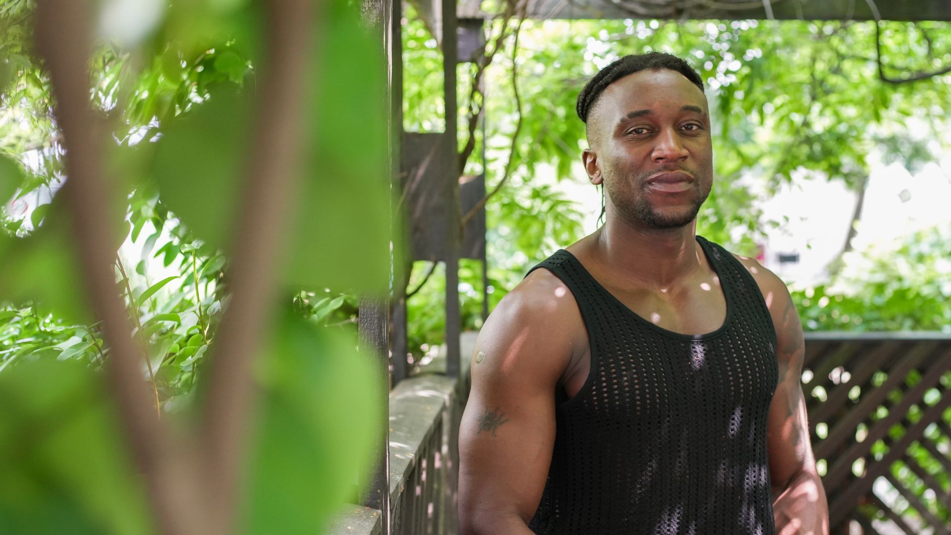 Khalif standing on a wooden deck surrounded by greenery, wearing a black mesh tank top, symbolizing his commitment to rebuilding life through fitness after homelessness and mental health challenges.