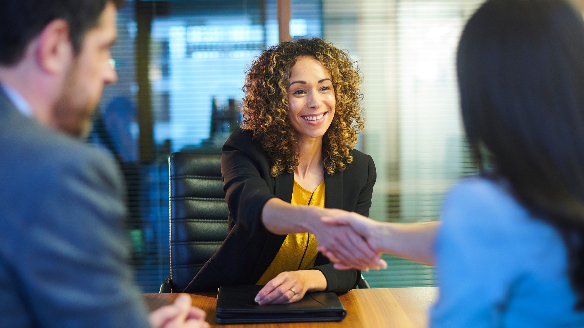 Smiling female job candidate in a black blazer and yellow top shaking hands with an interviewer across a desk.