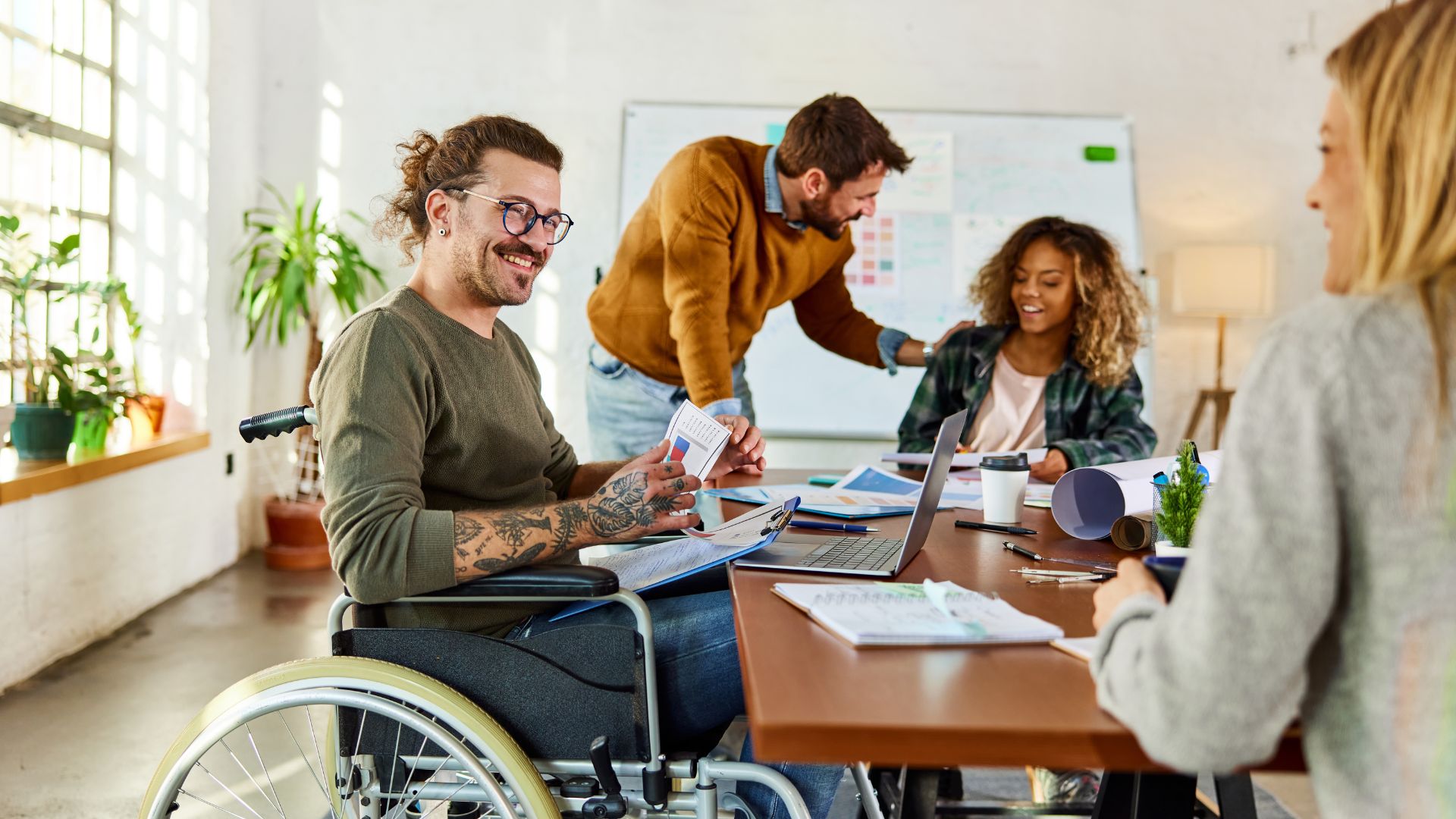 A man using a wheelchair speaking with colleagues in a meeting room, with three coworkers listening and smiling around a table.