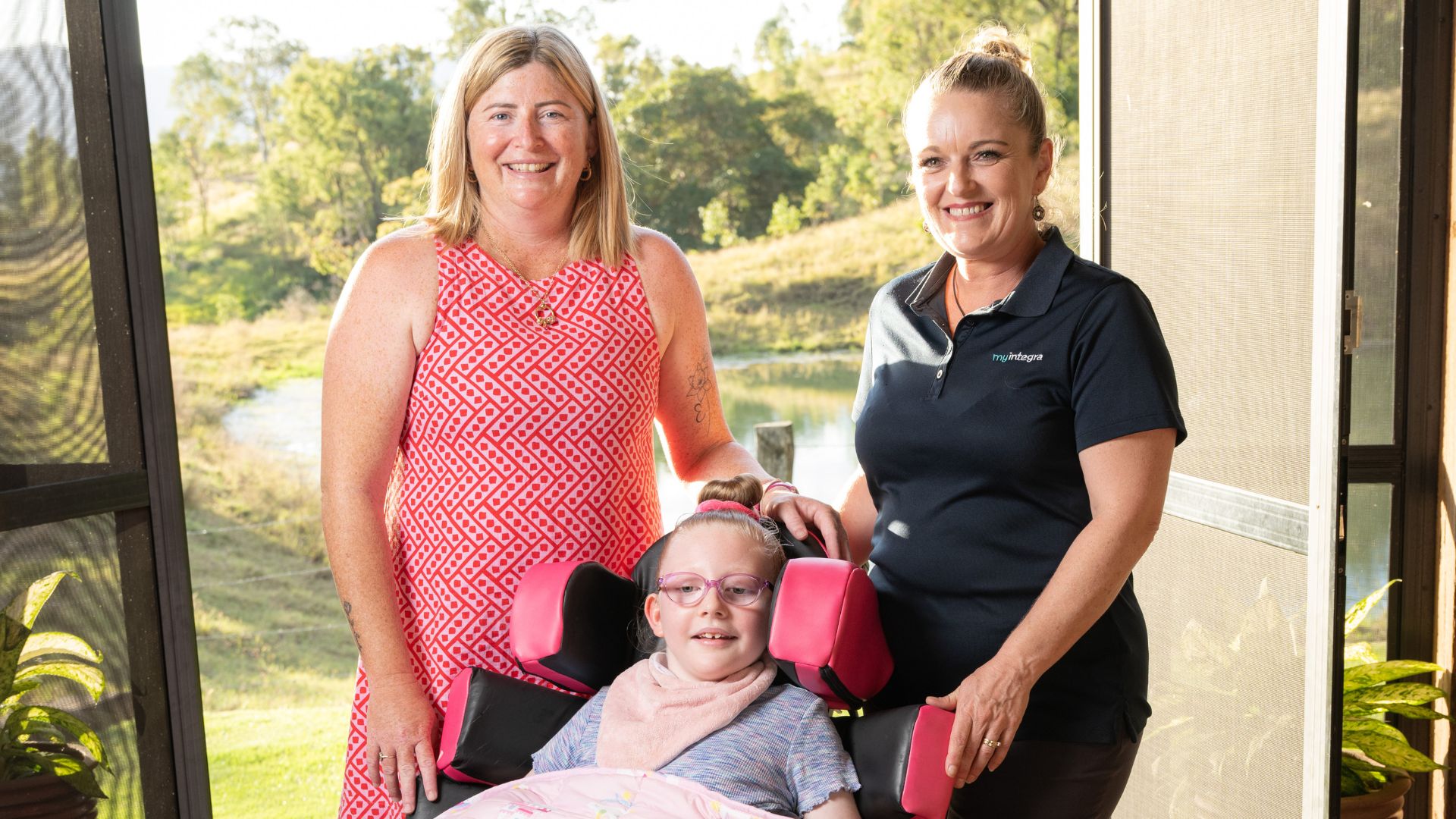 Jessica stands beside Emily, who is seated in a specialized wheelchair with pink head supports, alongside a MyIntegra support coordinator. They are outdoors on a sunny day with greenery and a pond in the background, symbolising family and disability support. 