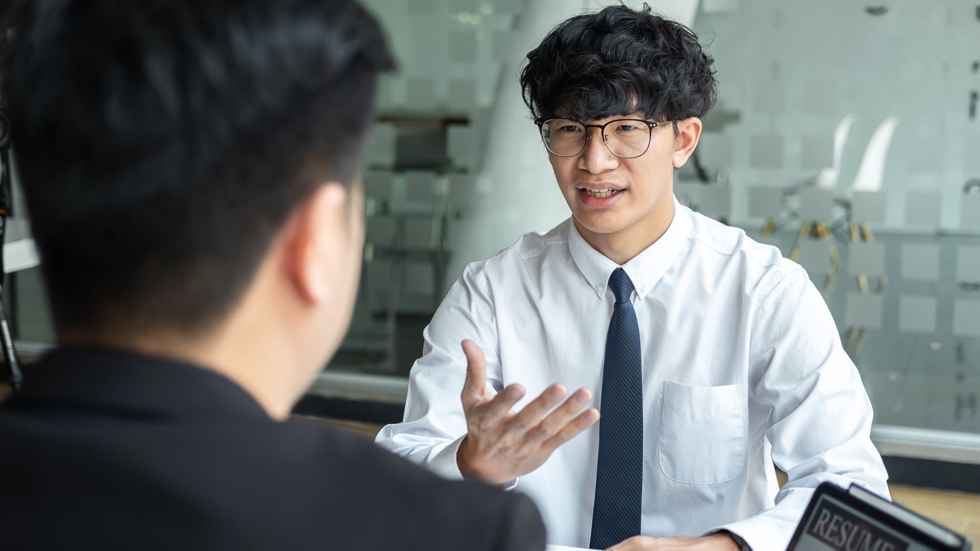 A young man in a white dress shirt and tie speaks animatedly during a job interview. He wears glasses and gestures with one hand while a recruiter, seen from behind, listens.