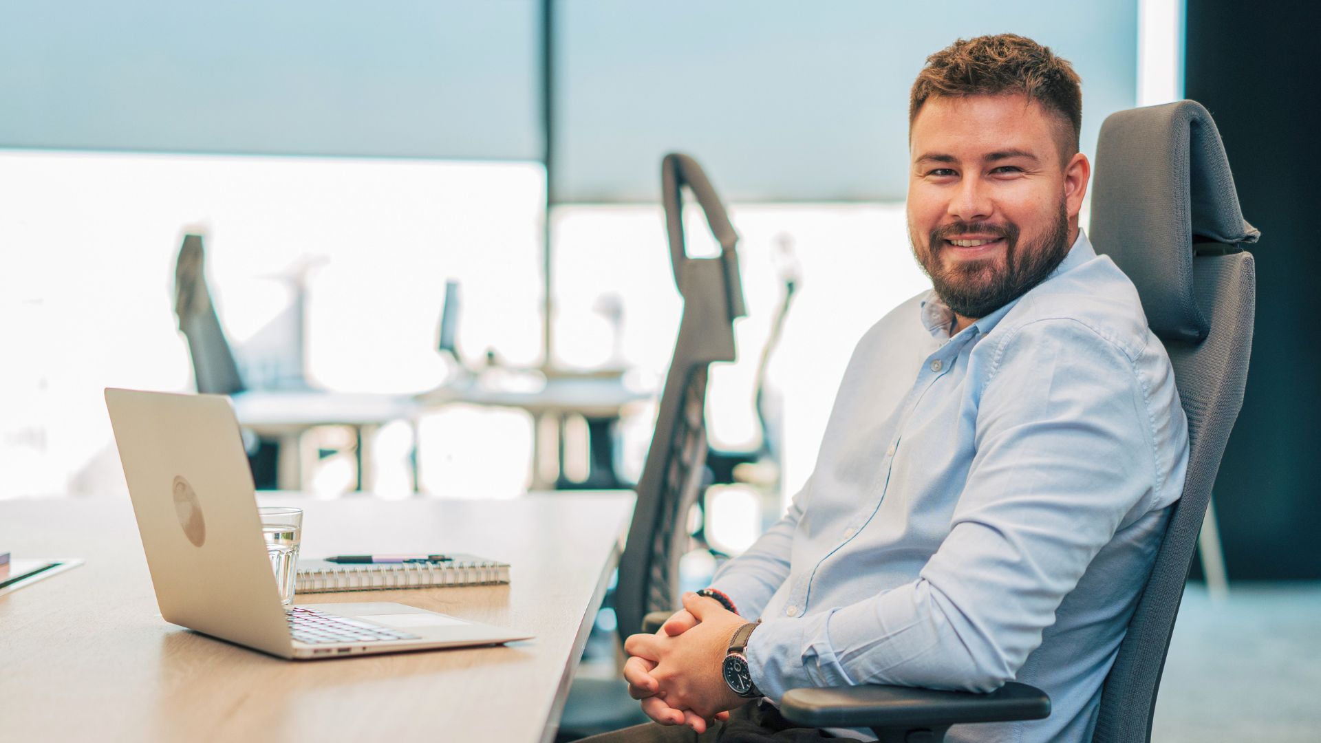 Smiling man sitting at a desk in an office, working on a laptop in a comfortable, ergonomic workspace.