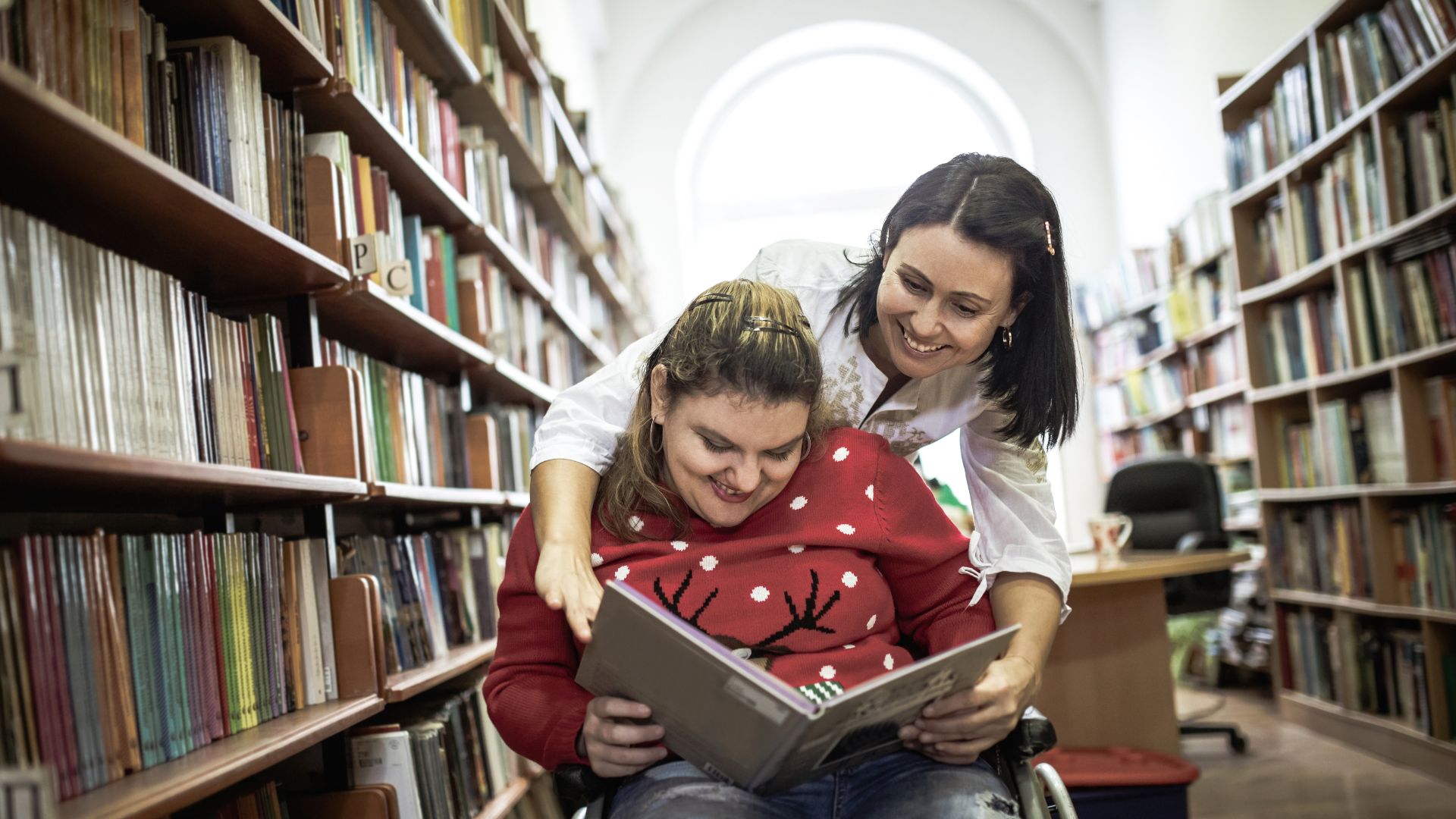 Young woman smiles reading a book with the support of a helper as she sits in a wheelchair in a library