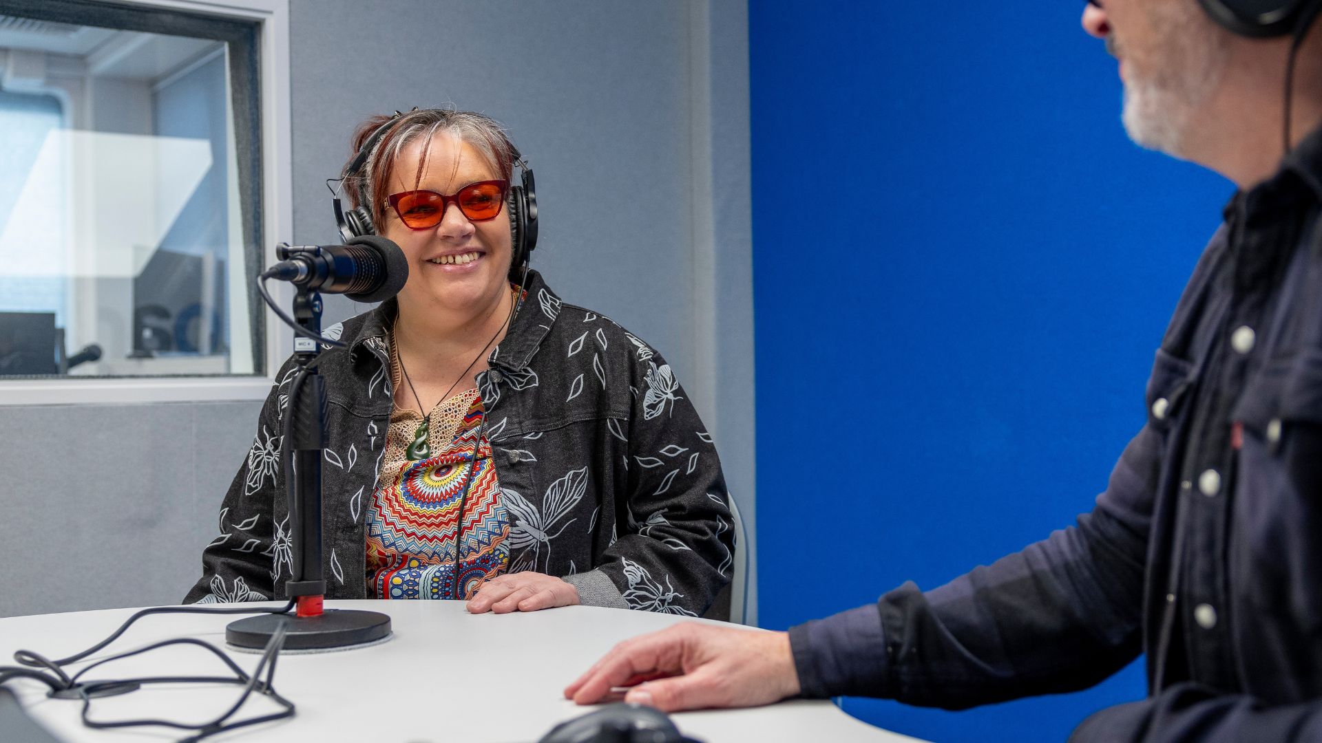 A woman sits at a radio microphone and smiles at a person at a control deck