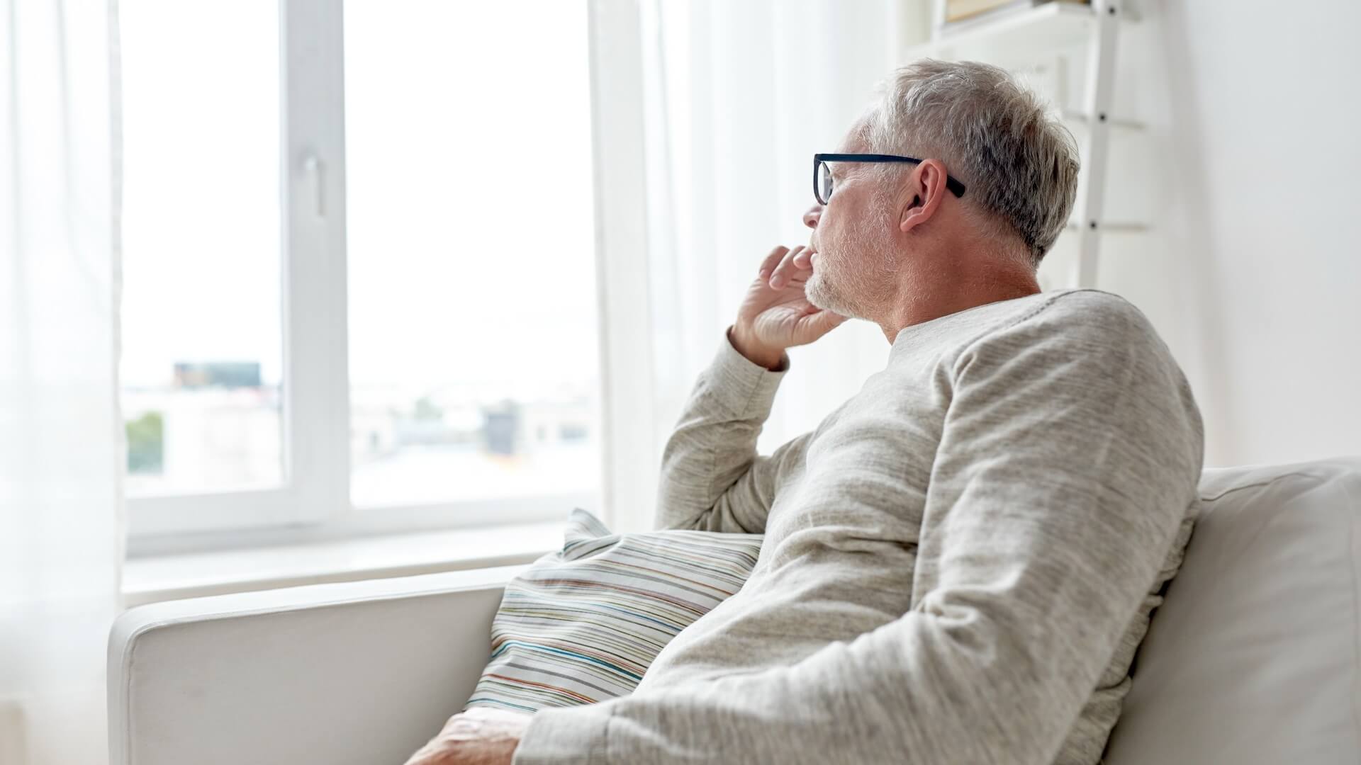 Older man sitting on a couch and looking out the window thoughtfully, resting his hand on his face