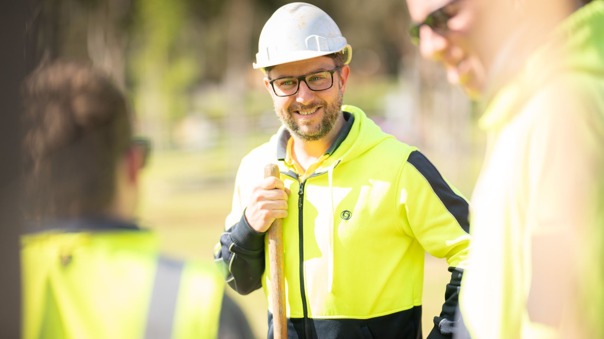 A construction worker wearing a hard hat and high-visibility jacket smiles while holding a measuring tape, with two colleagues partially visible in the foreground.