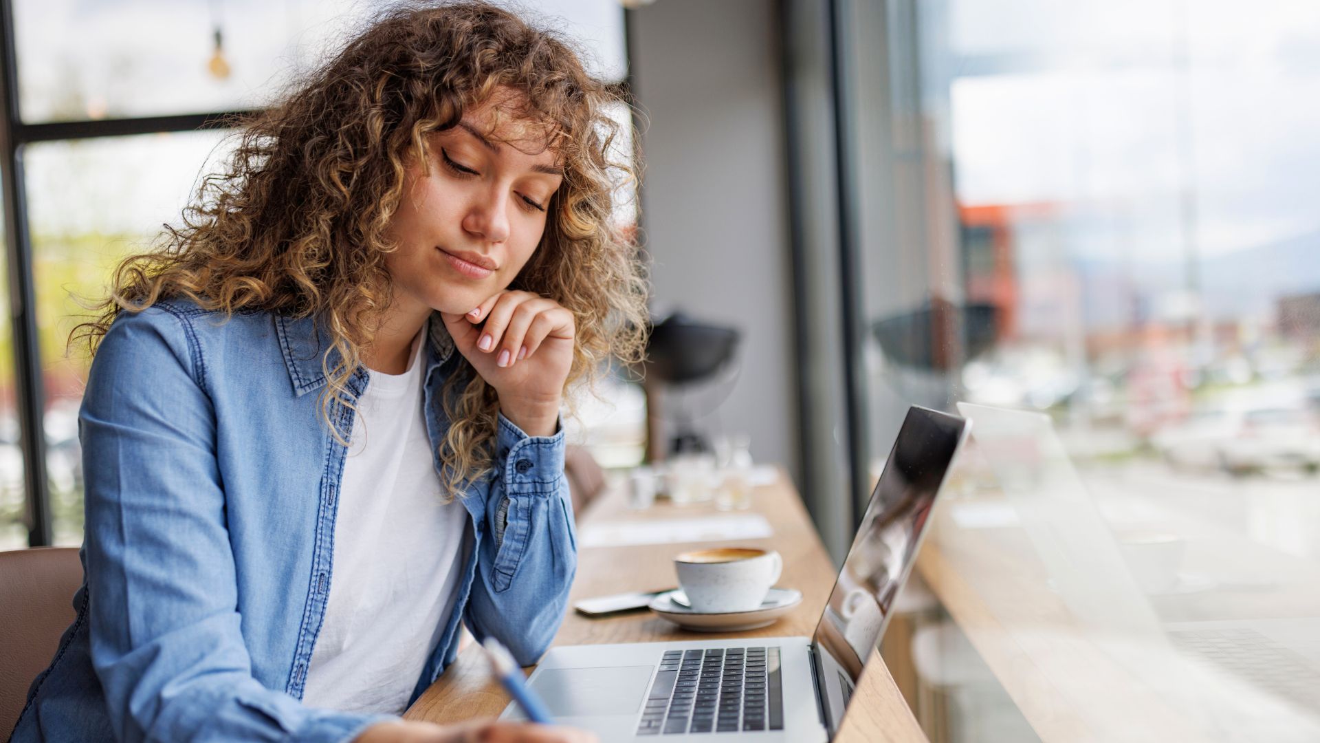 Woman writing notes in a notebook while working on a laptop at a café table.