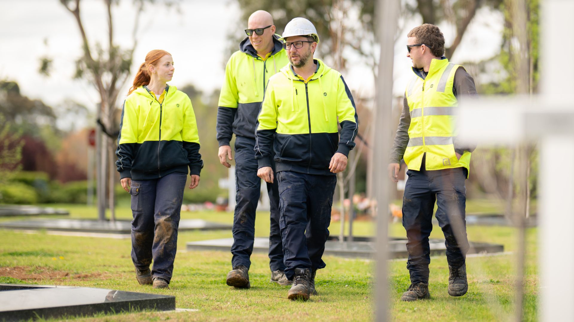 Four workers wearing high-visibility jackets walk through a park, engaged in discussion.