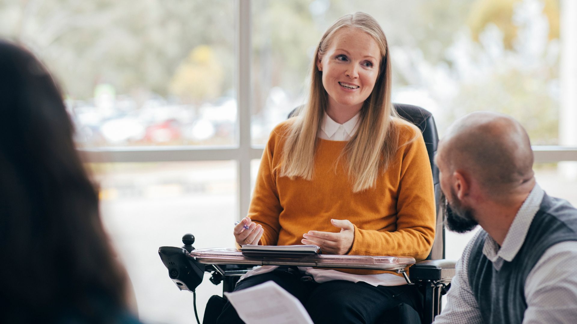 Person using a wheelchair speaking with two people during a meeting in an office setting.
