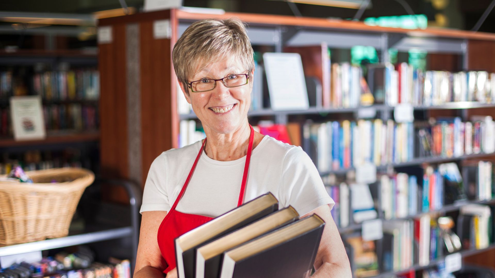 A woman in a red apron smiles carrying books in a library