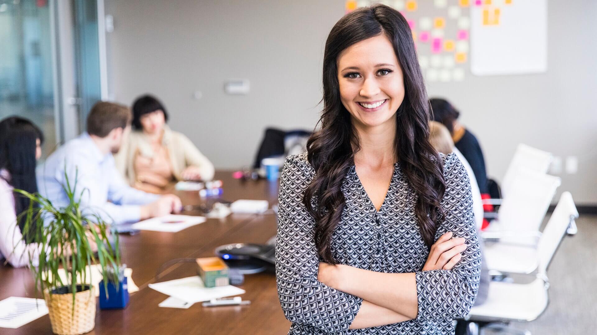 A smiling woman stands in front of her colleagues working in a boardroom