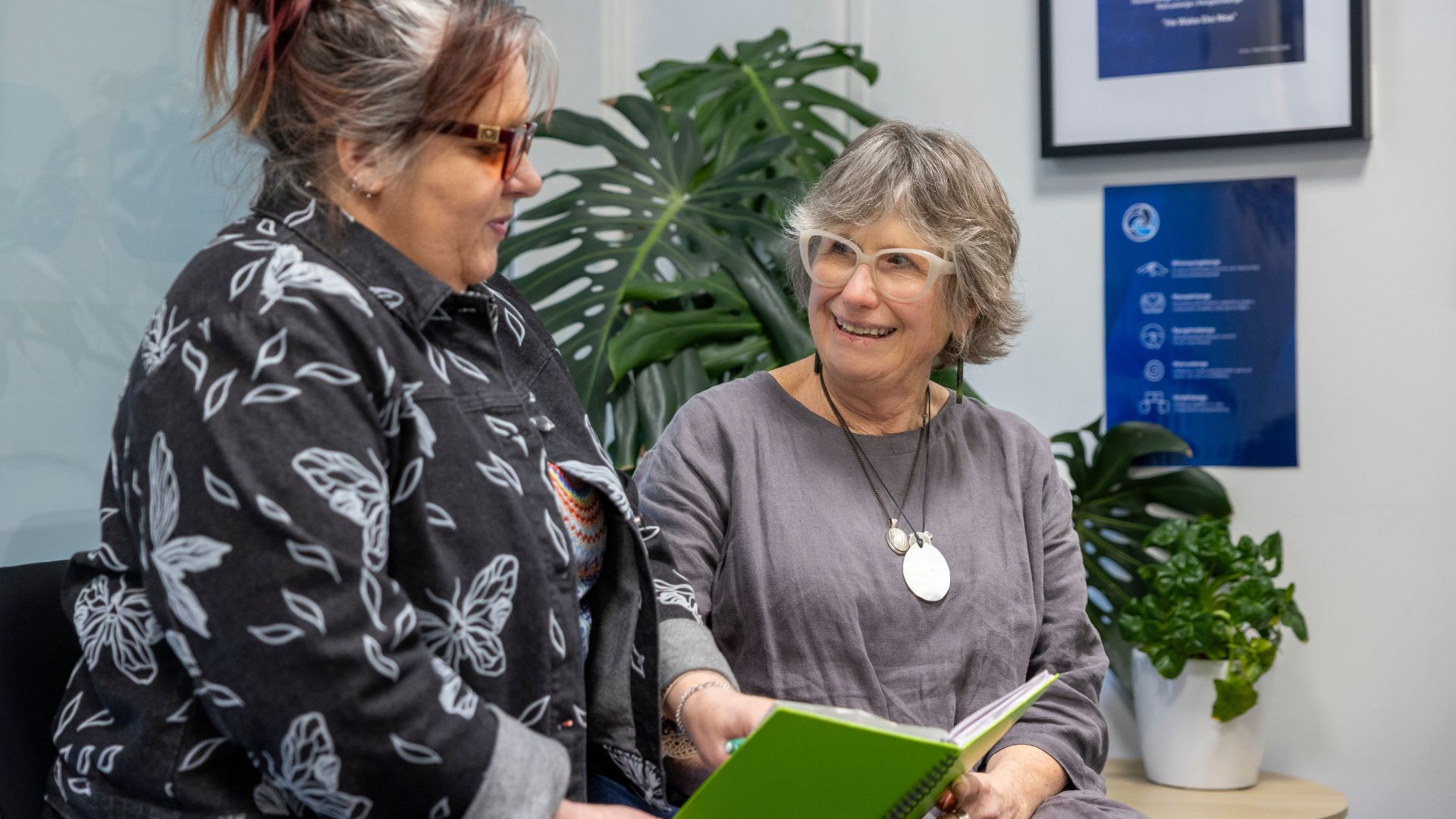 Two women smile and talk while looking and holding a green book.