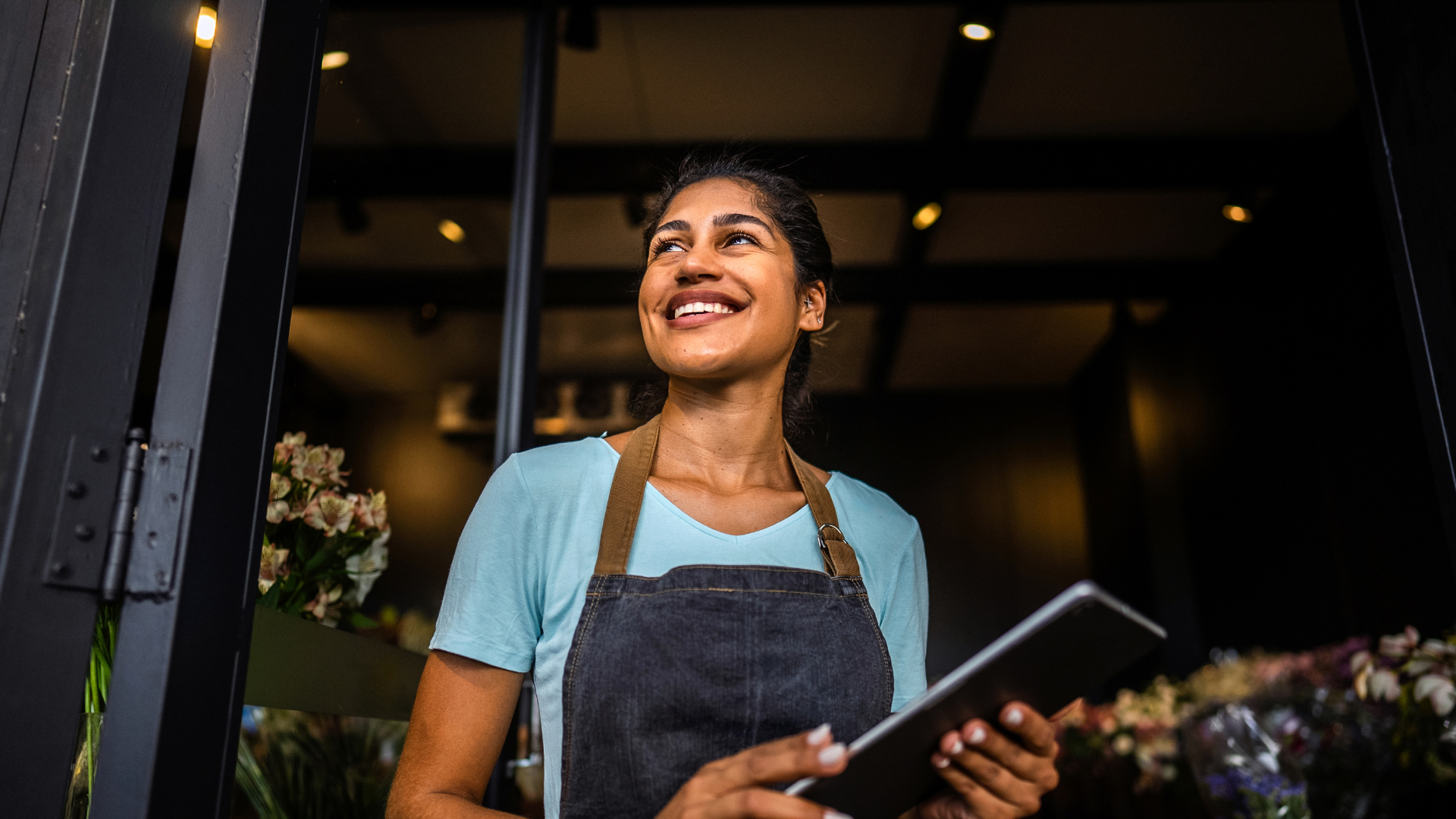 Female business owner smiling standing in the door of her shop, wearing apron, Self-Employment Assistance