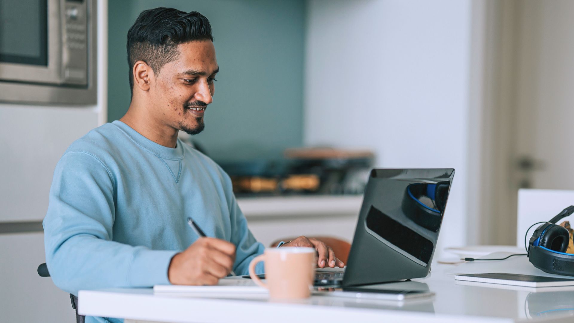 Person seated in a wheelchair using a laptop at a table, with a notebook and coffee nearby.