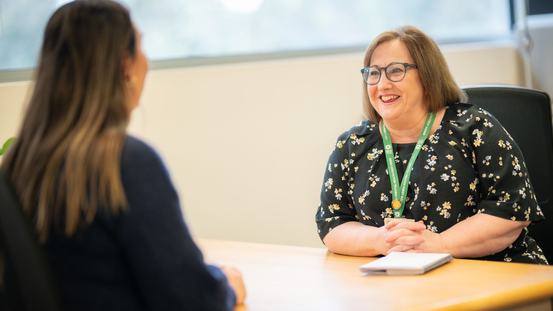 Two people are engaged in a conversation at a desk in a brightly lit office. One person is smiling broadly and wearing a lanyard with a badge, seated across from the other person whose back is facing the camera.