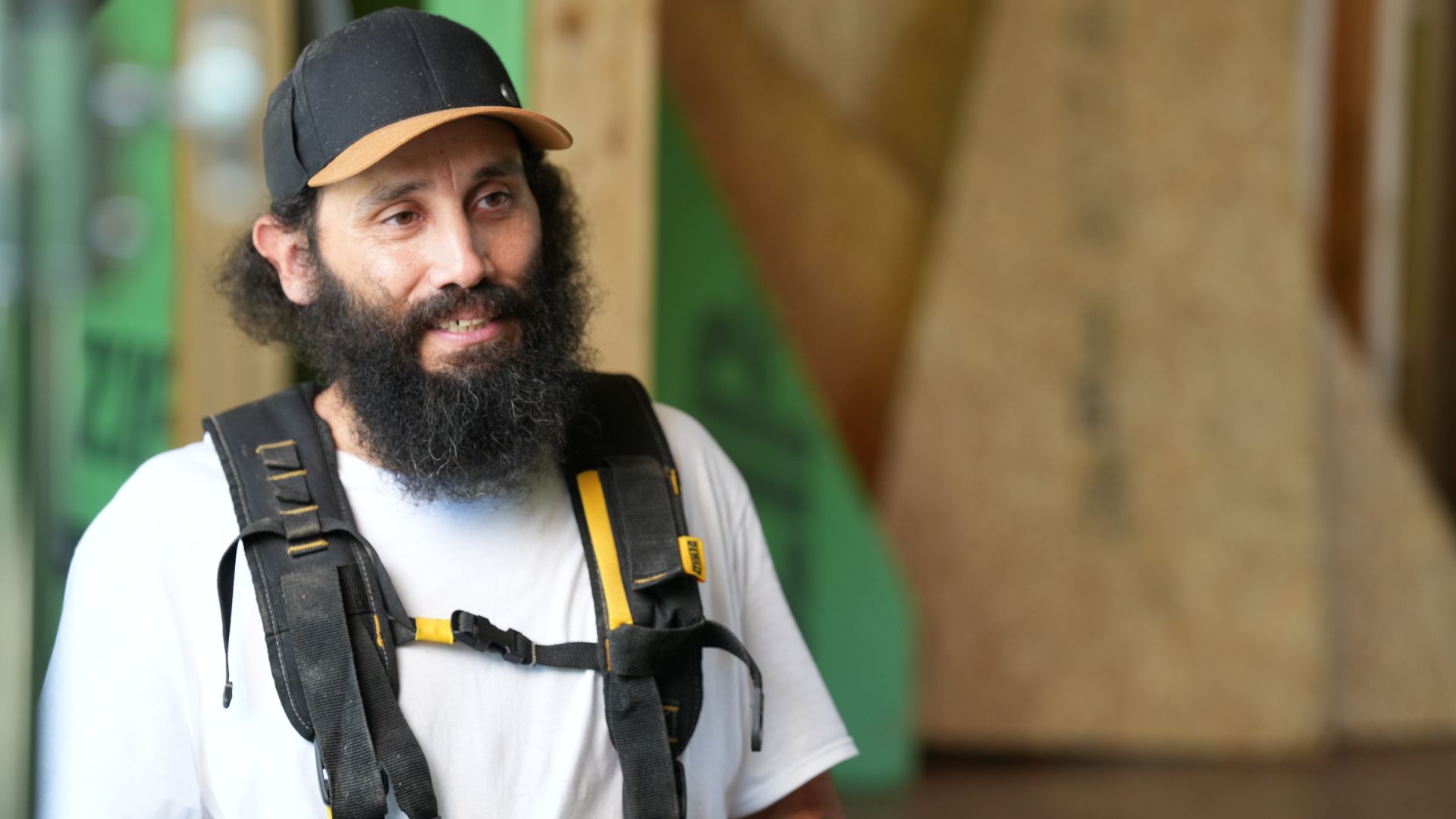 Person with a beard wearing a baseball cap and work overalls, smiling in a workshop setting.