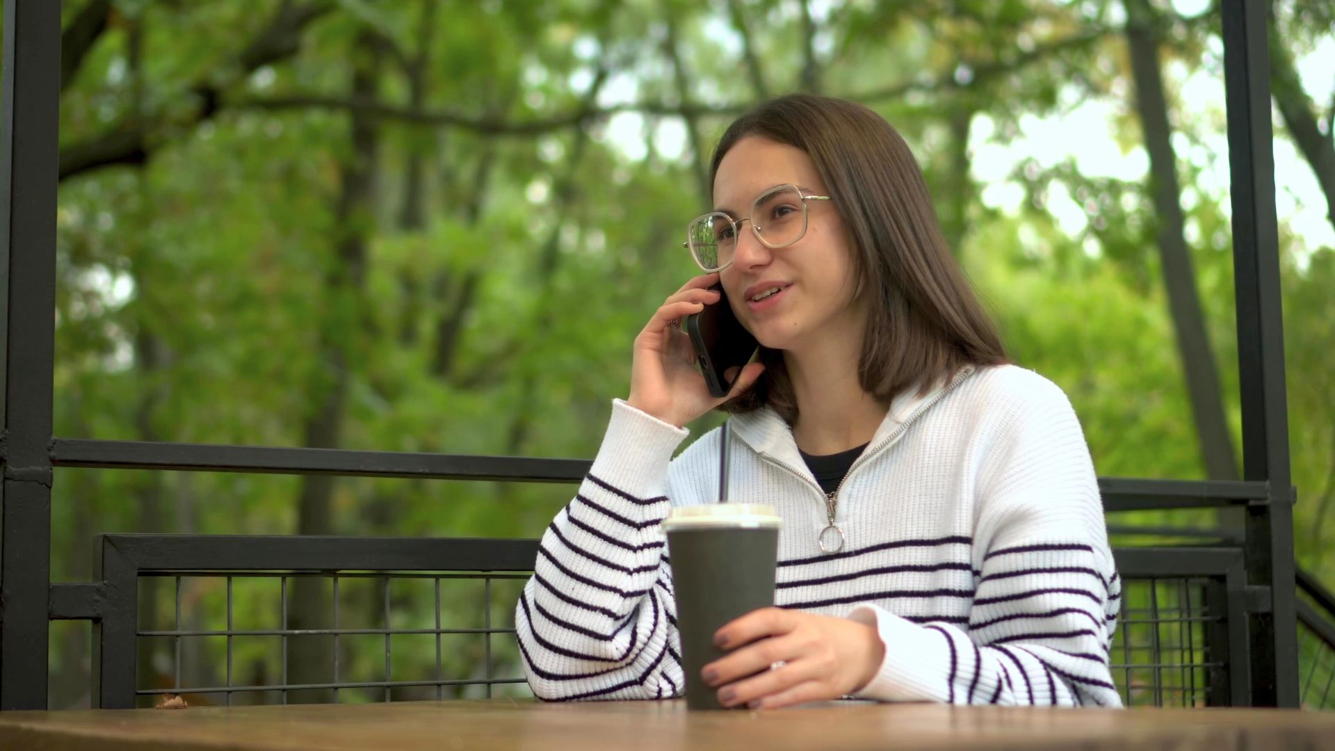 A young woman sitting at an outdoor café table, smiling while talking on her mobile phone, with a takeaway coffee cup in front of her.
