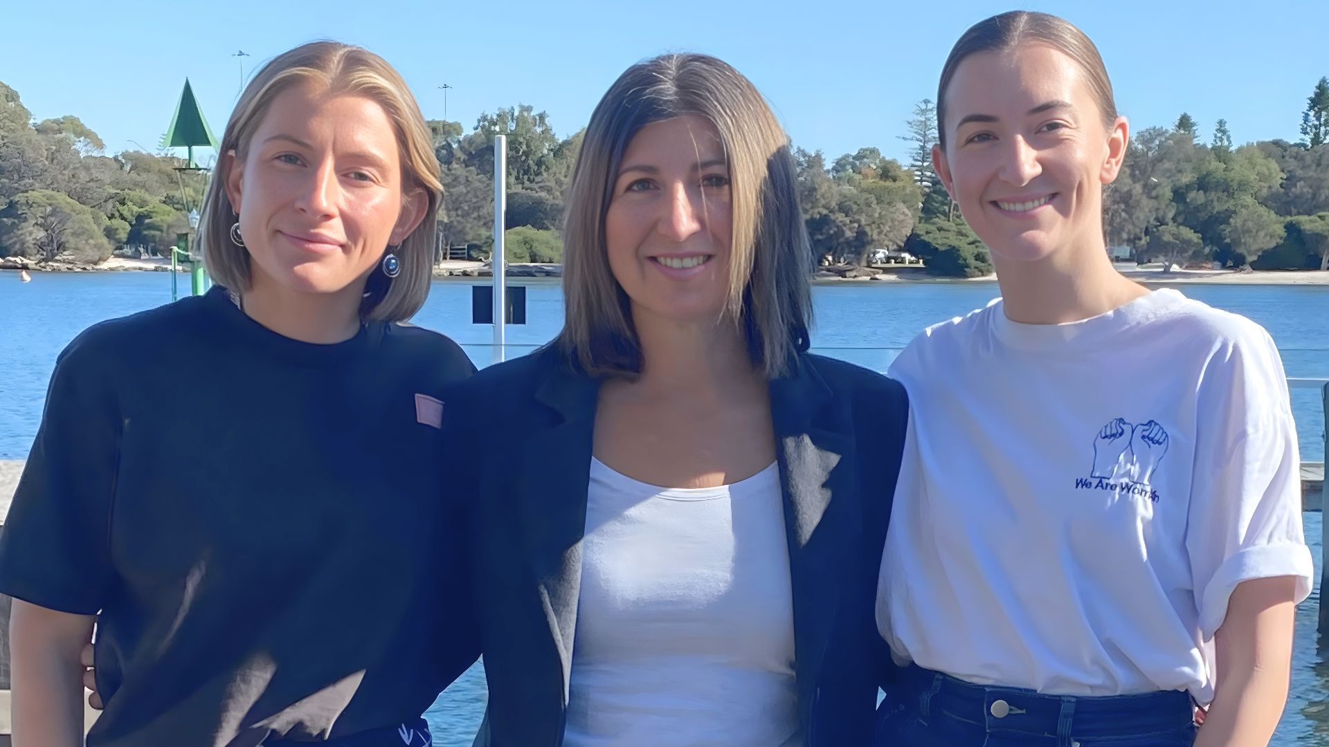 Self Employment Assistance participants Kate and Nicolette standing together outdoors near a waterfront with trees and clear skies in the background.