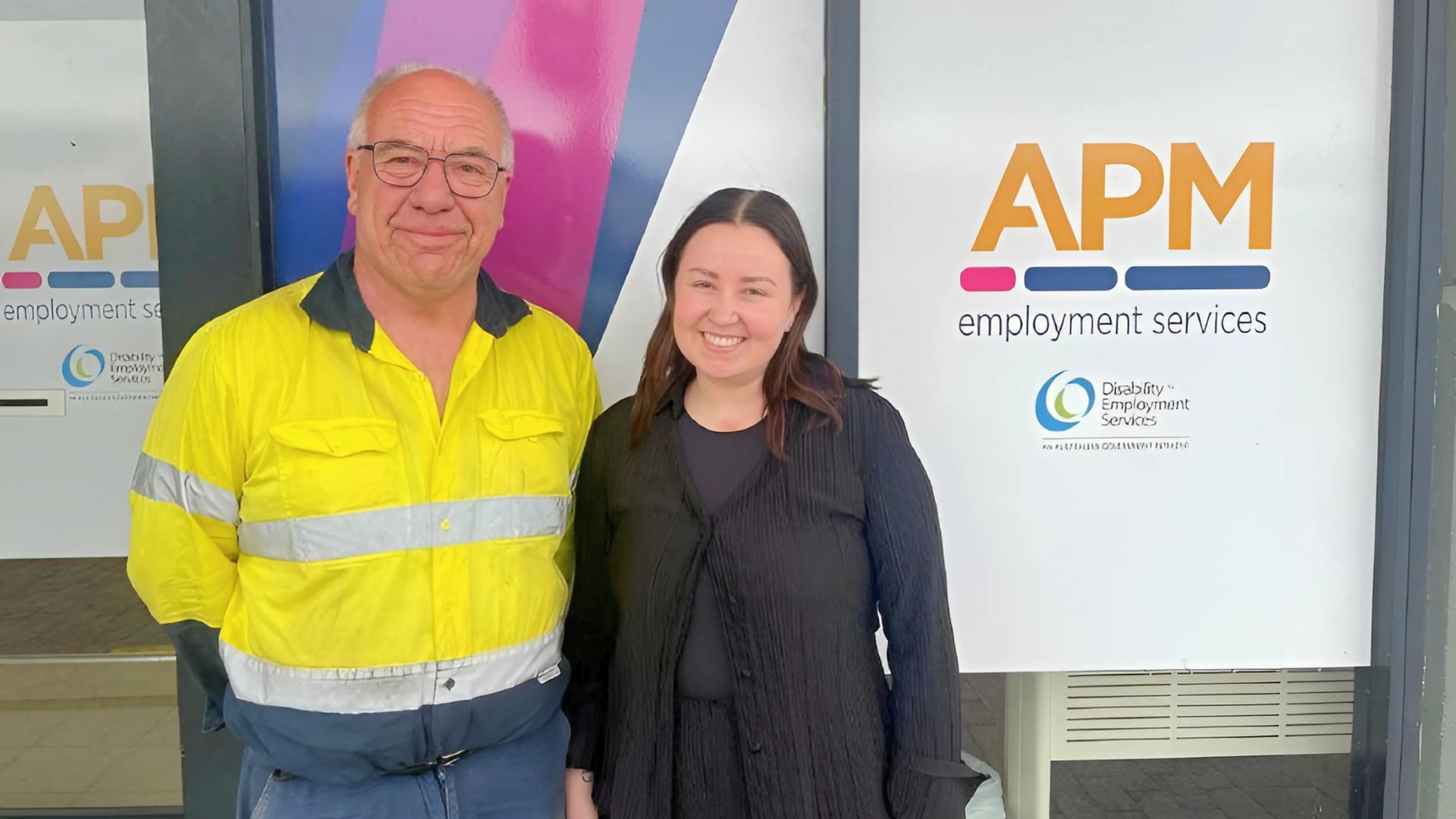 Daniel in a high vis jacket outside an APM office with employment consultant Crystal