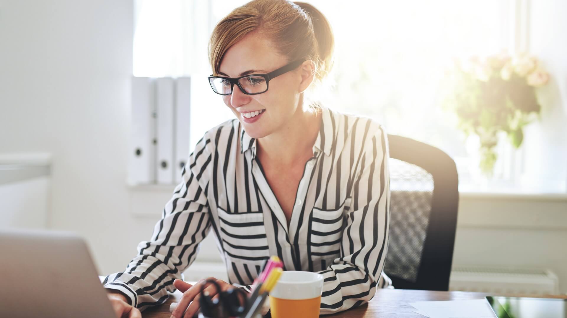Woman smiles while working on her laptop at a home office desk. The desk holds stationery, papers, a phone, and an orange coffee cup.