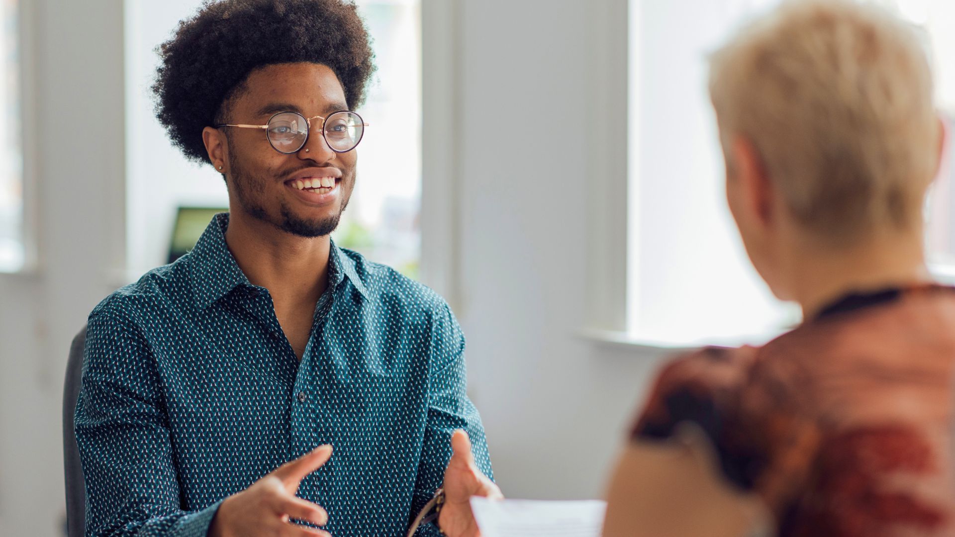 Young man wearing glasses smiling and speaking confidently during a job interview while holding his hands open in conversation, seated across from an interviewer reviewing a document.