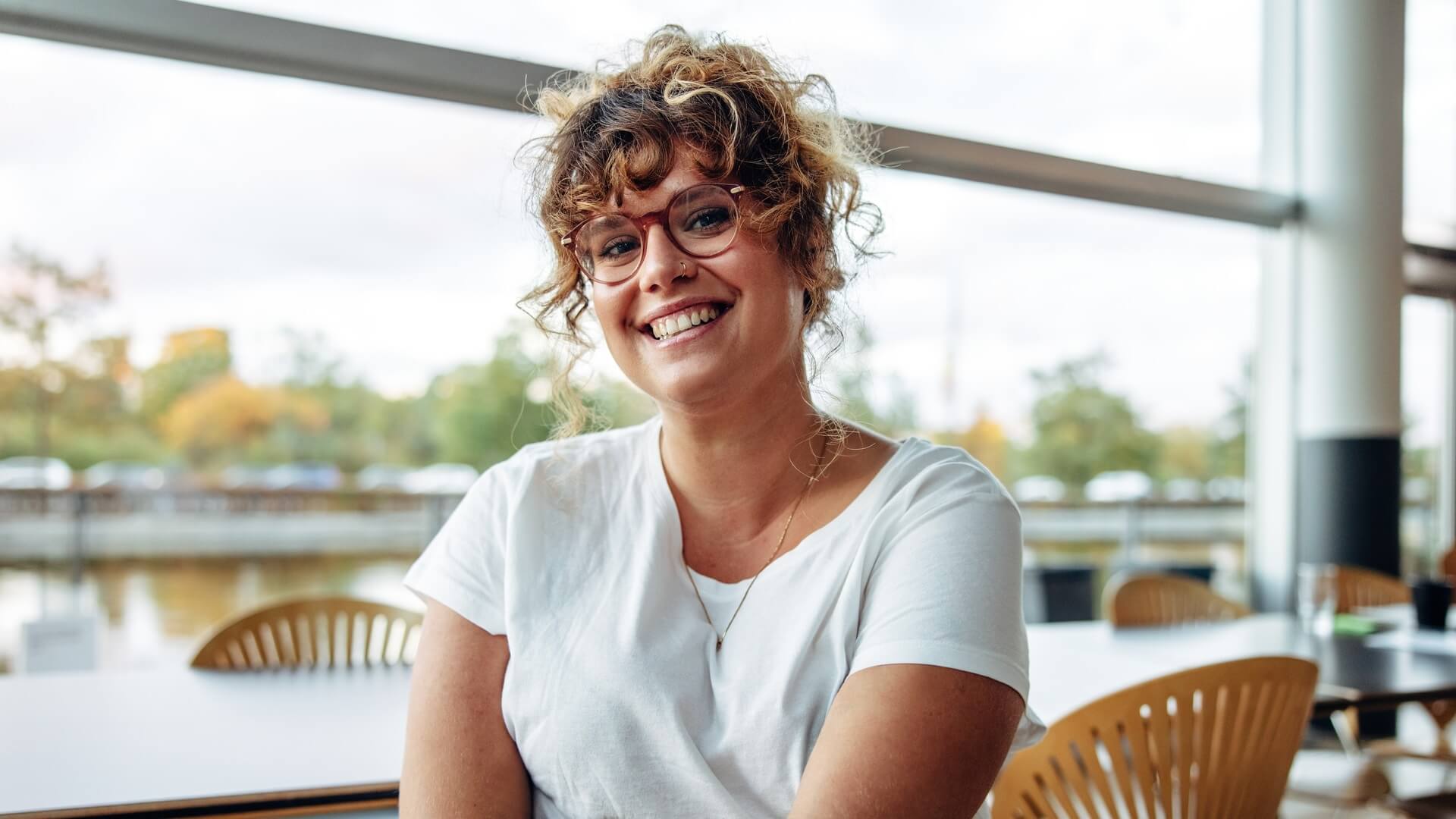Smiling woman with curly hair and glasses sits at a table near a large window, wearing a white T-shirt and looking relaxed.