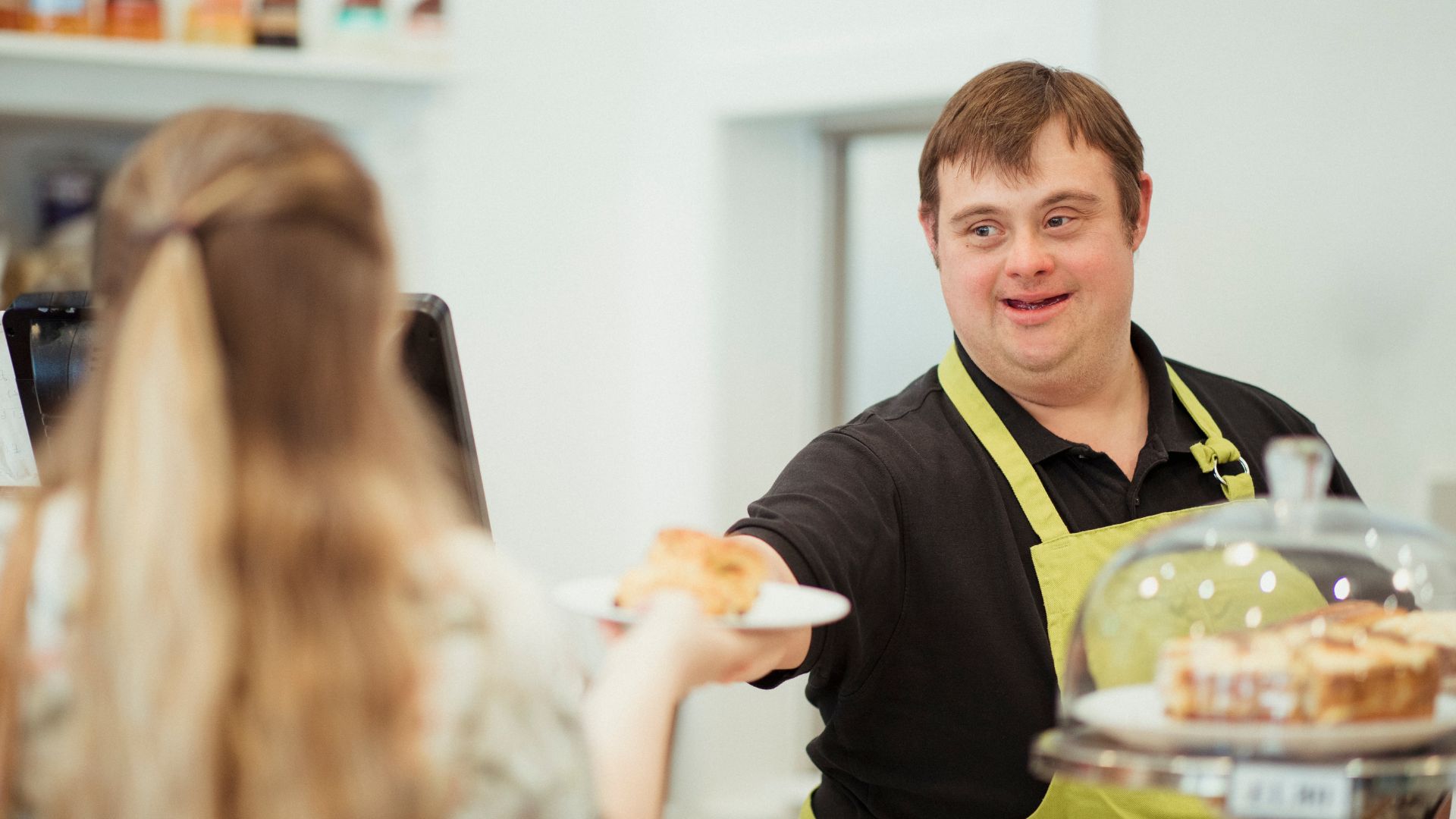 A café worker wearing an apron smiles while handing a plate of food to a customer across the counter, with baked goods and café equipment visible in the foreground.