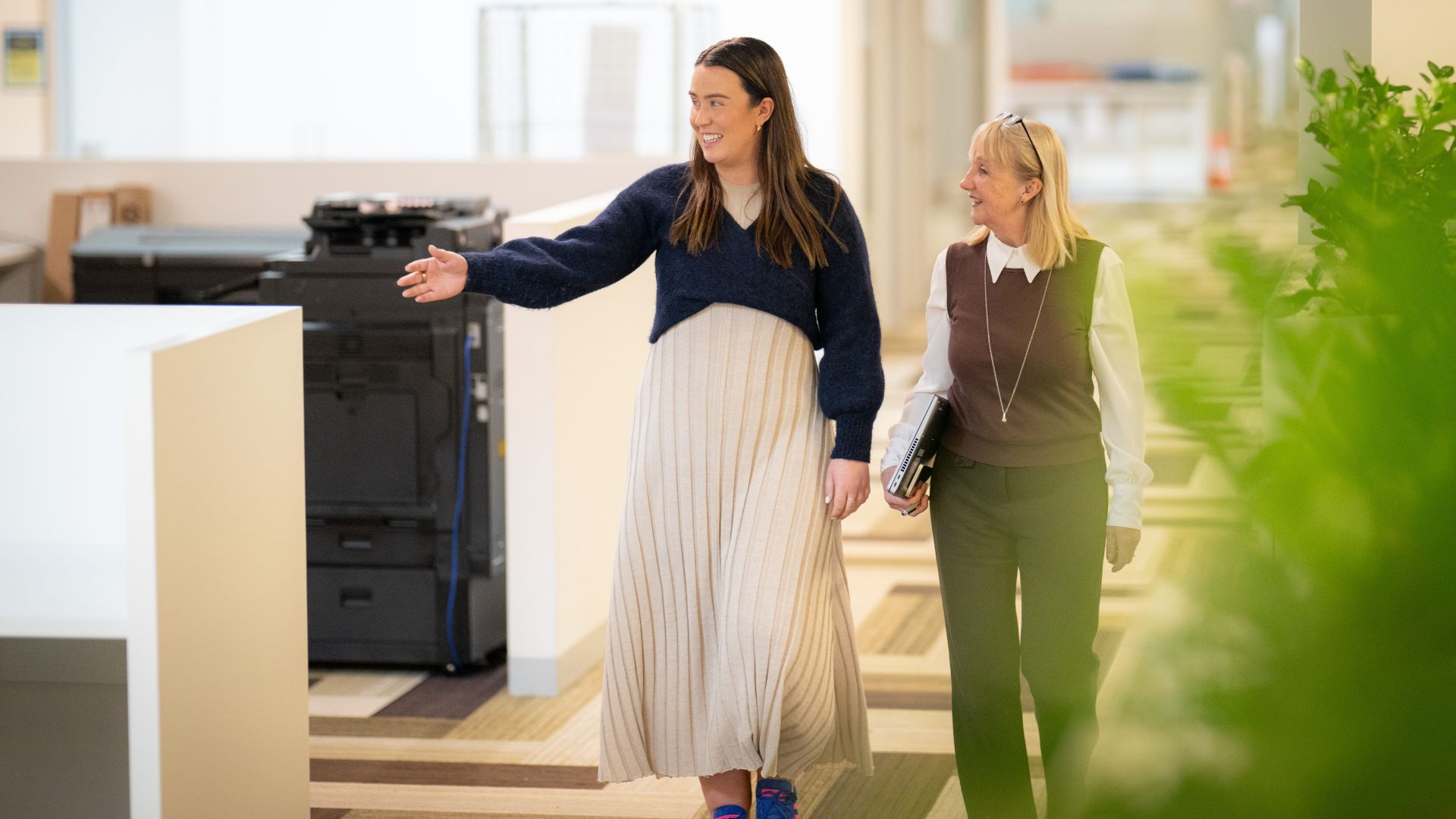 Two people walking and smiling in an office corridor, one is pointing at something out of view.