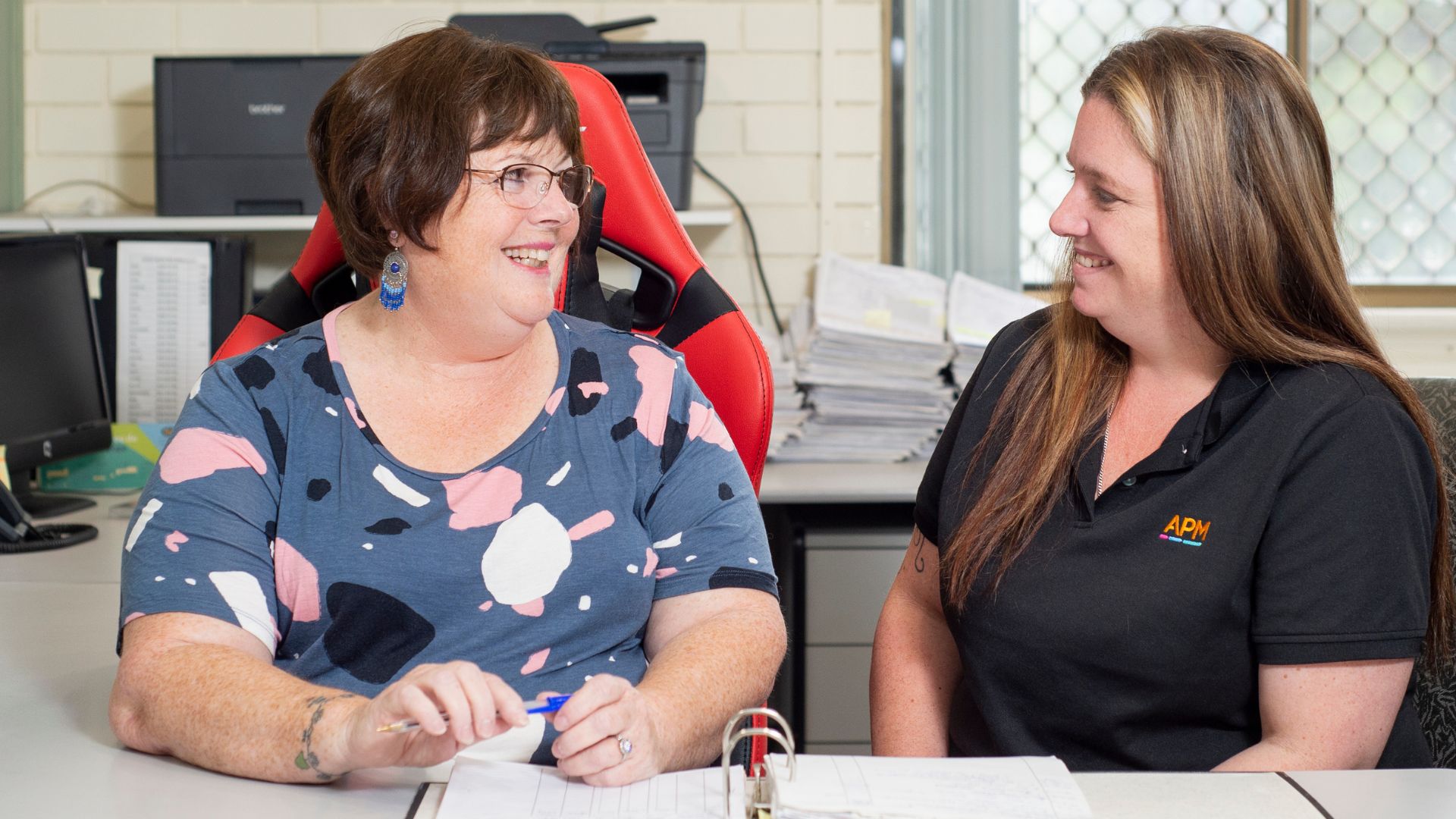 Woman speaking with an APM employment consultant at a desk, reviewing paperwork together in an office setting.